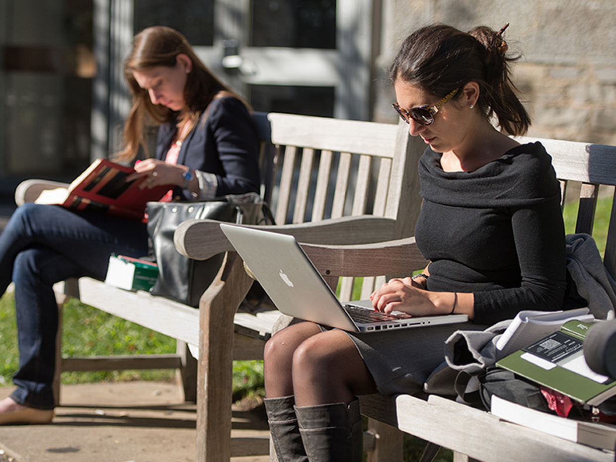 Two students on a bench using a laptop and reading.