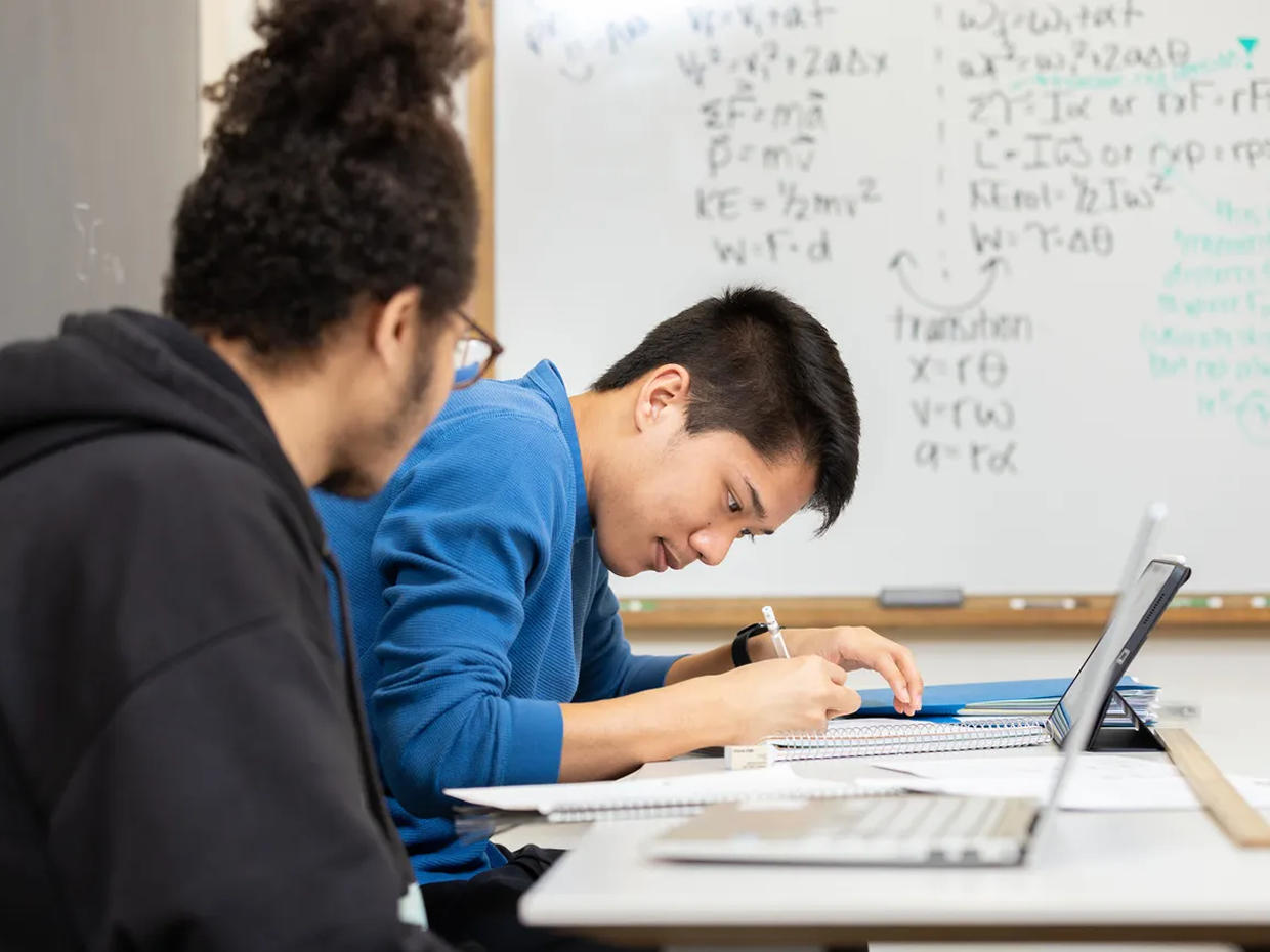 Students studying together at a desk with a whiteboard behind them.
